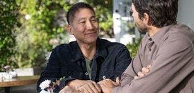 Two adult males smiling, seated, and facing towards each other in outdoor seating area