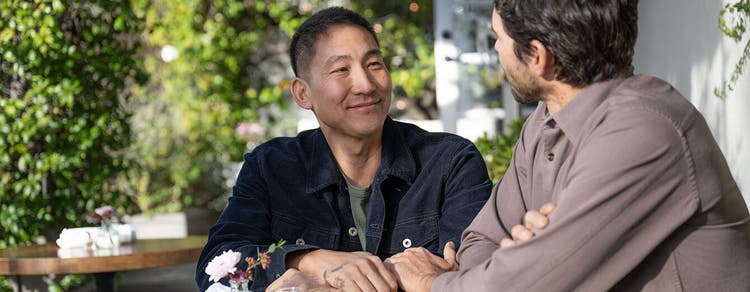 Two adult males smiling, seated, and facing towards each other in outdoor seating area