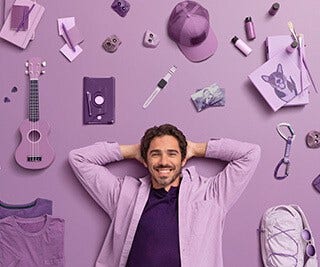 Smiling adult male with relaxed pose laying down on purple backdrop surrounded with various lifestyle items.