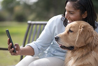 Smiling woman using her phone with her pet golden retriever sitting on a bench outdoors