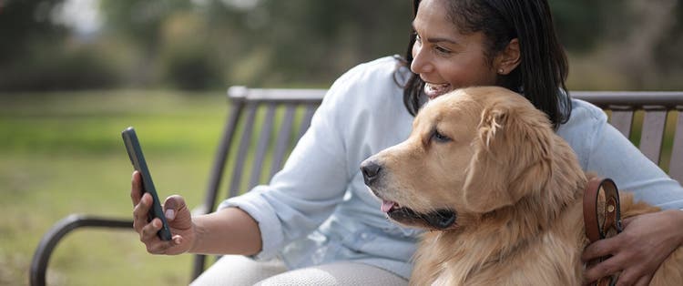 Smiling woman using her phone with her pet golden retriever sitting on a bench outdoors